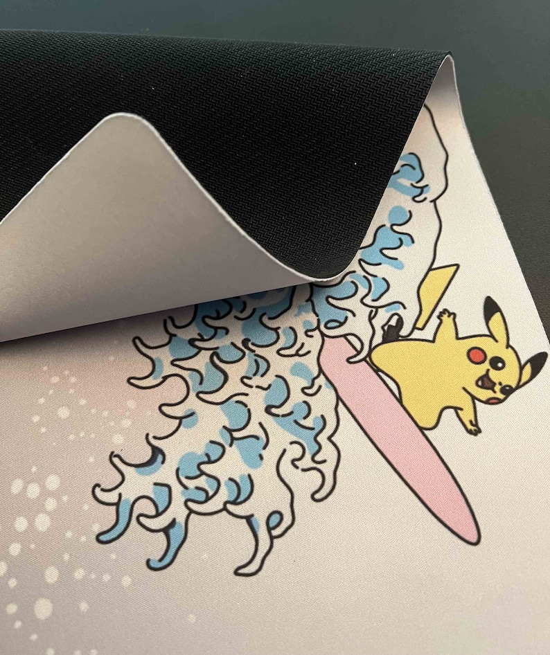 Op de afbeelding: Een witte muismat met een cartoon afbeelding van Pikachu die op een roze surfplank op een blauwe golf rijdt. De muismat heeft een zwarte rubberen achterkant.