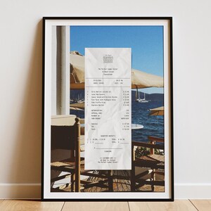 May include: A receipt from Summer House Restaurant, 30 Beach Street, Mykonos, Greece. The receipt is for a Dirty Martini, Salad Nicoise, Caesar Salad with Grilled Chicken,  and a side of truffle fries. The receipt is on a table with two chairs, a white umbrella, and a view of the ocean.