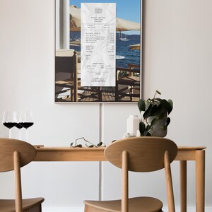 Peut inclure: Une impression encadr&eacute;e d'un re&ccedil;u d'un restaurant appel&eacute; "The Perfect Summer" avec une vue sur une mer bleue et des bateaux en arri&egrave;re-plan. Le re&ccedil;u est sur une table avec deux chaises. Il y a une plante dans un pot blanc sur une table en bois au premier plan.