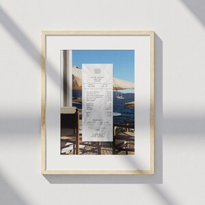 Peut inclure: Un re&ccedil;u du restaurant The Village Gourmet Kitchen en Gr&egrave;ce, avec vue sur l'oc&eacute;an et des bateaux en arri&egrave;re-plan. Le re&ccedil;u liste diff&eacute;rents plats et leurs prix, dont une salade grecque, des poulpes grill&eacute;s et une bouteille de vin. Le co&ucirc;t total du repas est de 45,50 euros.