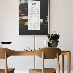 May include: A framed print depicting a blurred night street scene with yellow taxis and a crumpled NYC receipt. Below, a wooden table is set with two chairs, wine glasses, and a potted plant.