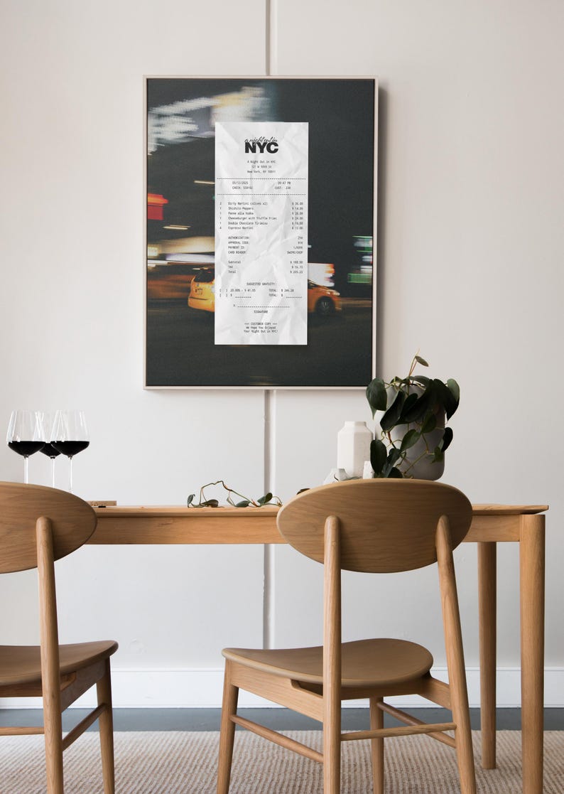 May include: A framed print depicting a blurred night street scene with yellow taxis and a crumpled NYC receipt. Below, a wooden table is set with two chairs, wine glasses, and a potted plant.