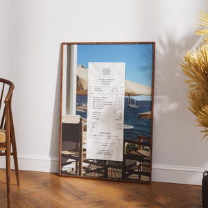 Peut inclure: Une photographie encadr&eacute;e d'un re&ccedil;u d'un restaurant appel&eacute; "The Pretty Sunset Tavern" avec une vue sur une &eacute;tendue d'eau bleue avec des bateaux et un parasol blanc au premier plan.