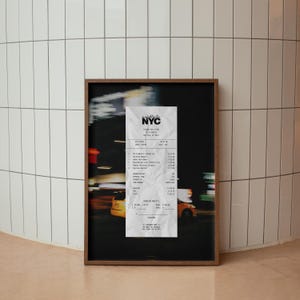 May include: A framed print showing a crumpled NYC receipt against a blurred cityscape. The receipt has black text on white paper, and the frame is dark wood. The background features yellow taxis and street lights.