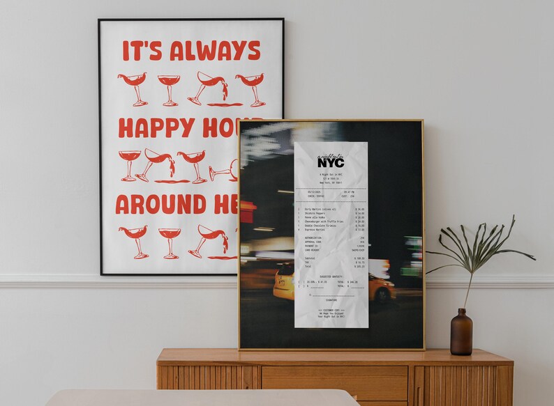 May include: Two framed art prints. One has red text saying "IT'S ALWAYS HAPPY HOUR AROUND HERE" with cocktail illustrations. The other is a photo of a NYC receipt.