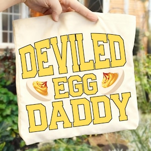 Peut inclure: Sac fourre-tout crème avec l'inscription "DEVILED EGG DADDY" en lettres jaunes, accompagné d'illustrations d'œufs mimosa. Le sac est tenu par une main.