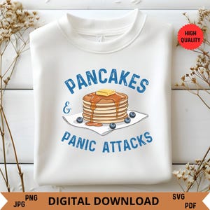 May include: White sweatshirt with blue text that reads "Pancakes & Panic Attacks". A cartoon image of a stack of pancakes with blueberries and syrup is above the text.