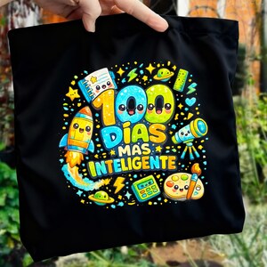 May include: Black tote bag with a vibrant cartoon design. The design includes the text "100 DÍAS MÁS INTELIGENTE" and illustrations of a rocket, telescope, calculator, and other school-related items. The bag is held by a hand.