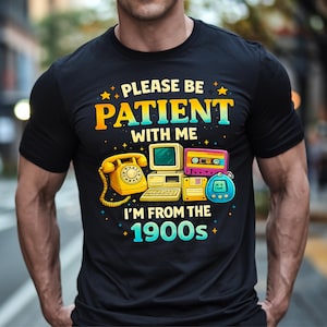 Peut inclure: T-shirt noir avec un motif coloré représentant un téléphone vintage, un ordinateur, une cassette et un jeu portable. Le texte dit : « PLEASE BE PATIENT WITH ME I'M FROM THE 1900s ».