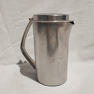 Puede incluir: Una jarra de plata con tapa abatible y asa. La jarra es de metal y tiene un acabado liso y brillante.