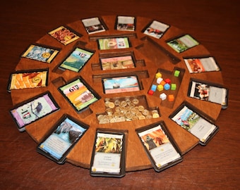 May include: A circular wooden game board with slots for cards and a compartment for gold coins. The board is made of dark wood and has a number of cards in the slots, including cards with numbers and pictures of buildings. There are also several gold coins in the compartment.