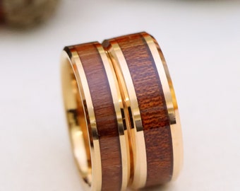 May include: Two wedding rings with a gold band and a dark wood inlay. The rings are set against a soft, neutral background, with blurred acorns in the background. The rings are a symbol of commitment and love.