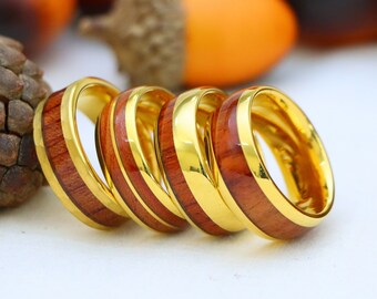 May include: A collection of gold-colored rings with a polished wood inlay. The rings are arranged in a row, showcasing the contrast between the warm wood and the shiny gold. The background features blurred fall-themed elements.