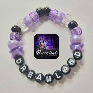 May include: Purple and black beaded bracelet with the word "DREAMLAND" spelled out in black beads. The bracelet also features star-shaped beads and a small image of a person in a forest.