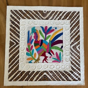 May include: A square decorative wall hanging featuring a central embroidered panel with a colorful bird and floral motif. The embroidery is surrounded by a white embossed border and an outer frame of brown and white woven geometric patterns.
