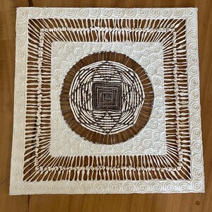 May include: A square wall art piece featuring a central circular woven design in dark brown and cream fibers. The center is surrounded by a cream-colored border with embossed spiral patterns and a fringe of vertical cream threads connecting to the edge.