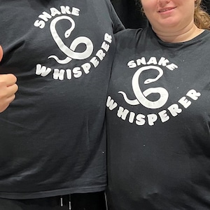 May include: Two people wearing black t-shirts with a white snake design and the text "Snake Whisperer". Both are giving a thumbs up.
