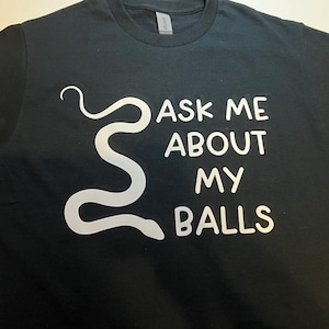 May include: Black t-shirt with white text and a stylized snake graphic. The text reads "ASK ME ABOUT MY BALLS". The shirt has a crew neck and short sleeves.