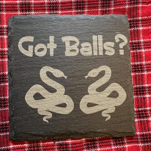 May include: Black slate coaster with the text "Got Balls?" in white lettering. Two gray snake silhouettes are on either side of the text.