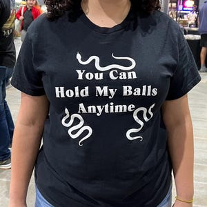 May include: Black t-shirt with the text "You Can Hold My Balls Anytime" in white, accompanied by snake illustrations. The shirt is a crew neck style.