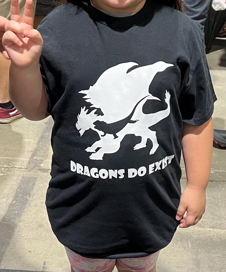 May include: A black t-shirt with a white silhouette of a dragon and the text "DRAGONS DO EXIST".
