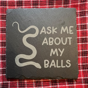 May include: A square, dark gray slate coaster with the phrase "ASK ME ABOUT MY BALLS" in a light gray font. A stylized snake design is on the left side of the text. The coaster is on a red and black plaid surface.