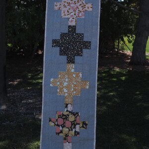May include: A long, rectangular table runner with a patchwork design. The runner is made of five different fabrics, each with a different pattern. The fabrics are arranged in a vertical pattern, with the top and bottom fabrics being pink with floral patterns, the middle fabric being a brown and white geometric pattern, and the two fabrics in between being black with white speckles and a brown and yellow floral pattern.