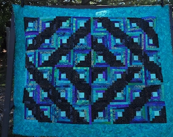 May include: A quilt with a log cabin pattern in shades of blue, green, and black. The quilt is made of fabric scraps and has a turquoise border.