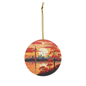 May include: A round ceramic ornament featuring a cityscape silhouette of Toronto, Canada, with the CN Tower in the center. The background is a sunset with a full moon and the text "TORONTO" is visible in the upper right corner. The ornament is decorated with fall foliage in shades of orange, red, and yellow.