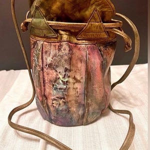 Peut inclure: Un sac seau avec fermeture à cordon, présentant une finition texturée multicolore dans des tons de rose, violet, vert et marron. Le sac possède un bord supérieur structuré avec des accents triangulaires et une longue bandoulière fine.