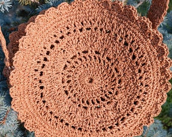 Può includere: Una borsa rotonda all'uncinetto marrone con un bordo smerlato e una lunga tracolla. Il design della borsa presenta un motivo a spirale che si irradia dal centro. La borsa è realizzata in un materiale tessuto e viene mostrata all'aperto su uno sfondo di aghi di pino blu-verdi.