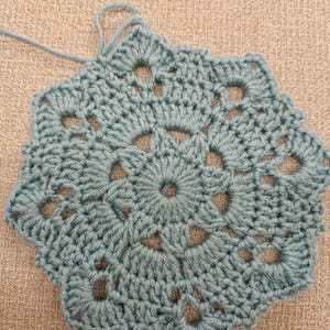 May include: A handmade, light teal crochet doily with a floral design. The doily features a central circular pattern surrounded by openwork and scalloped edges. The yarn is a soft, textured material.