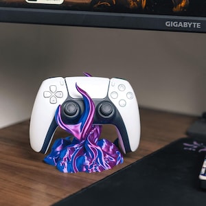 Pode incluir: Um comando PlayStation 5 branco e preto repousando sobre um suporte abstrato vibrante. O suporte apresenta um design em espiral com tons de azul, roxo e rosa. O comando está sobre uma superfície de madeira, com um monitor de computador ao fundo.