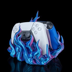 May include: A white and blue video game controller rests on a vibrant, flame-shaped stand. The stand is a mix of blue, purple, and pink hues, creating a dynamic visual effect. The controller has a sleek design, with buttons and a touchpad.