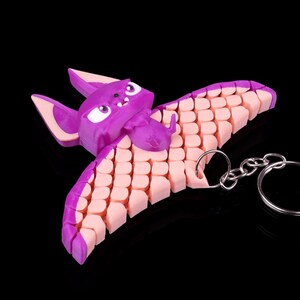 May include: A purple and pink bat-shaped keychain with a silver chain. The bat has a cartoon-like face with white eyes and a pink mouth. The wings are made of a series of small, pink, interlocking pieces.