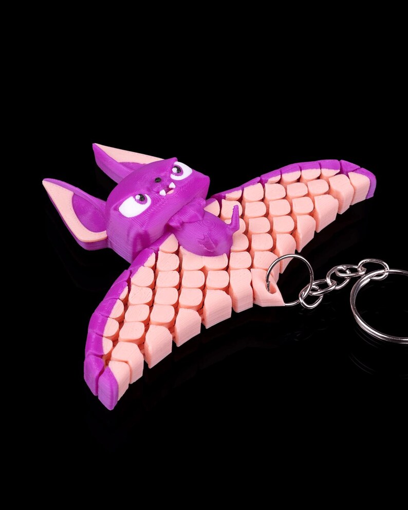 May include: A purple and pink bat-shaped keychain with a silver chain. The bat has a cartoon-like face with white eyes and a pink mouth. The wings are made of a series of small, pink, interlocking pieces.
