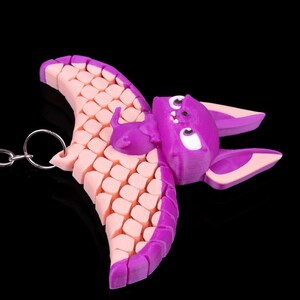 May include: A purple and pink bat keychain with a textured wing design. The bat has white eyes and a small, pink mouth.