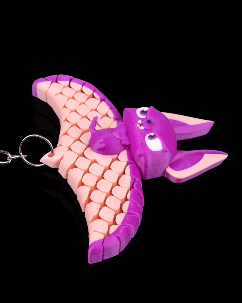 May include: A purple and pink bat keychain with a textured wing design. The bat has white eyes and a small, pink mouth.