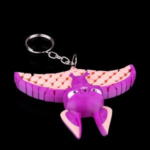 May include: A purple bat-shaped keychain with pink wings and a key ring. The bat has large eyes and a small nose. The wings are made of a textured material.