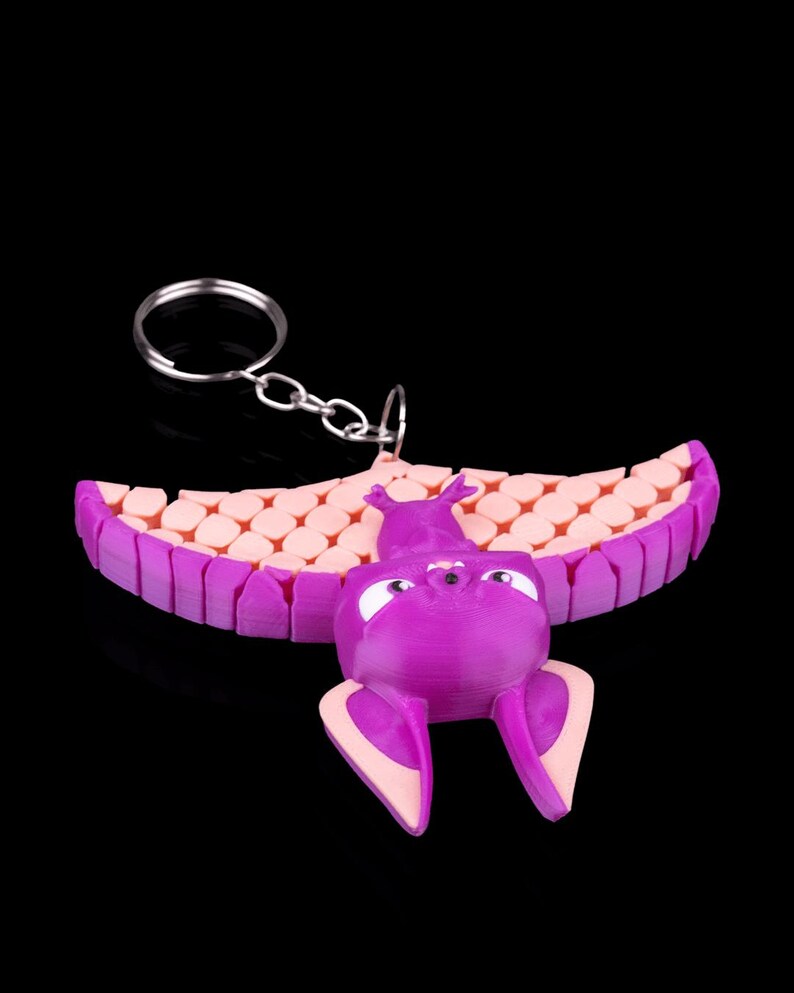 May include: A purple bat-shaped keychain with pink wings and a key ring. The bat has large eyes and a small nose. The wings are made of a textured material.