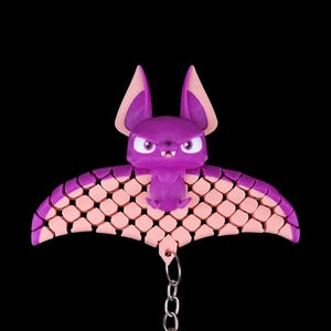 May include: A purple bat keychain with pink wings. The bat has a cute, cartoon-like face with angry eyes and a small mouth. The wings are made of a textured material that resembles scales. The keychain is attached to a silver chain.