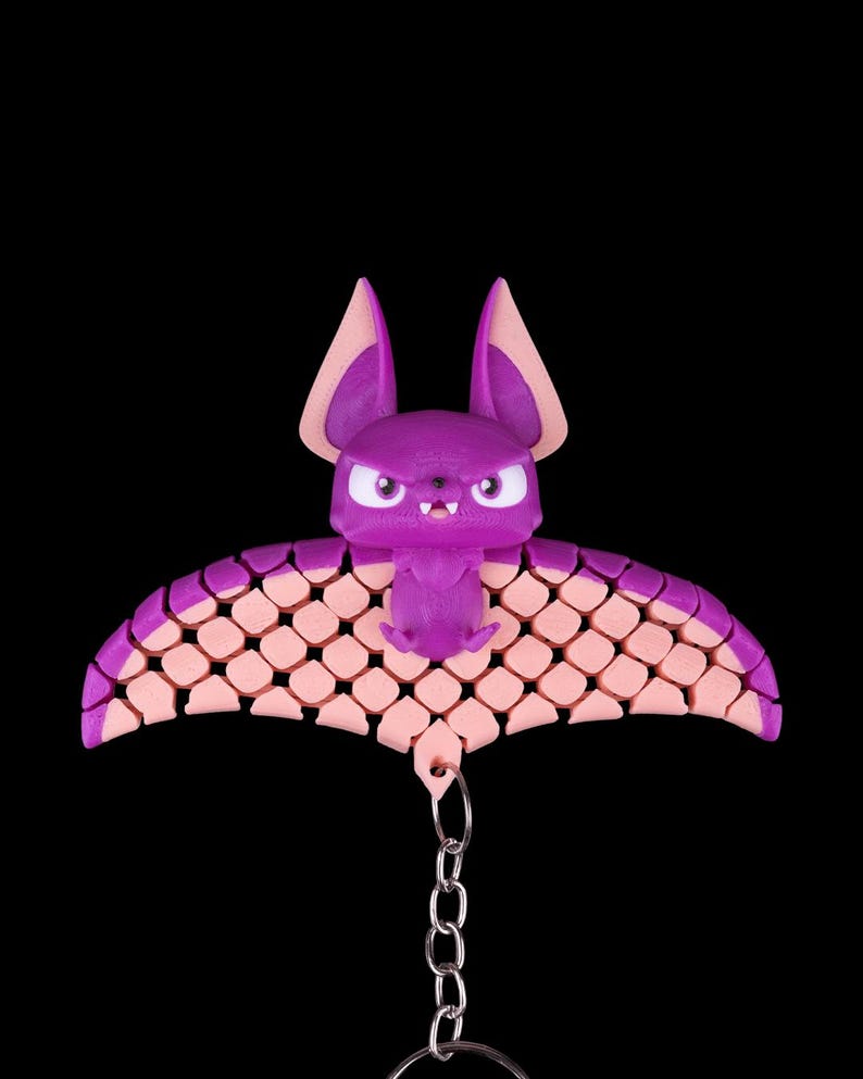 May include: A purple bat keychain with pink wings. The bat has a cute, cartoon-like face with angry eyes and a small mouth. The wings are made of a textured material that resembles scales. The keychain is attached to a silver chain.