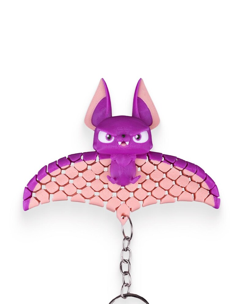 May include: A purple bat keychain with pink wings. The bat has a cute, cartoon-like face with angry eyes and a small mouth. The wings are made of a textured material that resembles scales. The keychain is attached to a silver chain.