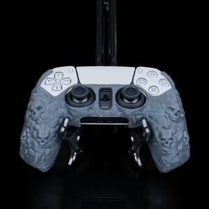 May include: A gray and white PlayStation 5 controller with a skull design. The controller is mounted on a black stand. The controller has black joysticks and buttons. The skull design is on the sides of the controller.