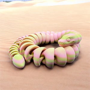 Puede incluir: Un juguete de serpiente de plástico segmentado de color rosa y verde. El juguete está enrollado en círculo sobre una superficie arenosa.