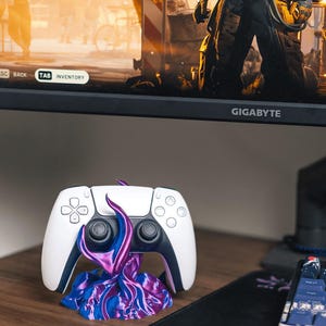 Może przedstawiać: Biało-czarny kontroler PlayStation 5 spoczywający na niestandardowym stojaku. Stojak ma wirujący wzór w odcieniach niebieskiego i fioletowego. Kontroler znajduje się na drewnianej powierzchni.