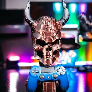Può includere: Un supporto per controller di gioco a forma di teschio color rame con corna argentate, che tiene in mano un controller PlayStation blu. Il supporto ha un design meccanico dettagliato. Il controller è appoggiato su una piccola base color rame.
