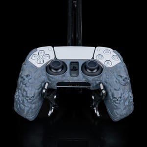 May include: A gray and white PlayStation 5 controller with a skull-patterned grip. The controller is on a black stand.