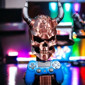 Puede incluir: Un cráneo de color cobre con cuernos, un mando de PlayStation azul y una base de color cobre. El cráneo está detallado con una superficie texturizada y ojos brillantes.