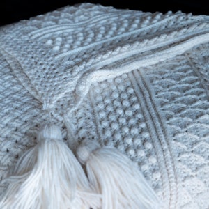 May include: Cream-colored knitted throw blanket with intricate patterns and two tassels. The blanket features various textures, including cable knit, bobbles, and diamond designs. The soft, cozy blanket is folded, showcasing its detailed craftsmanship.